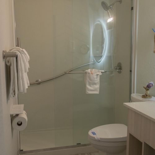 A bathroom with a glass shower door, round mirror, towel rack, toilet, and vanity with a sink on light-colored flooring.