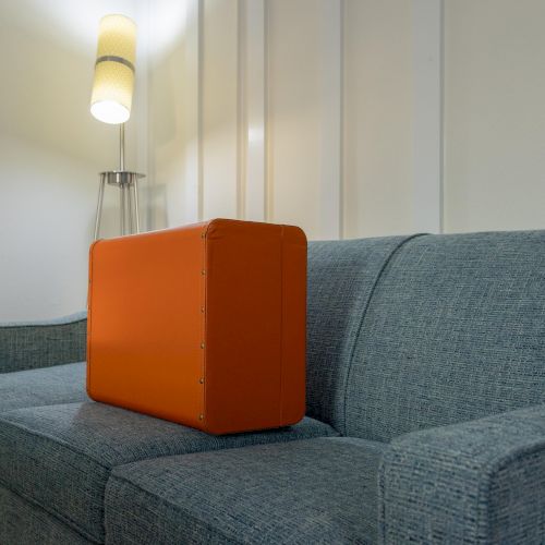 A blue sofa with an orange suitcase placed on it, beside a floor lamp, against a paneled wall in a living room setting.