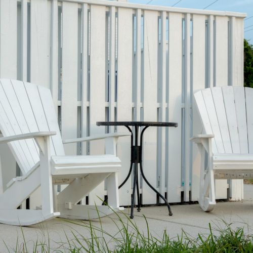 Two white Adirondack rocking chairs and a small black metal table are placed on a patio with a white wooden fence in the background.