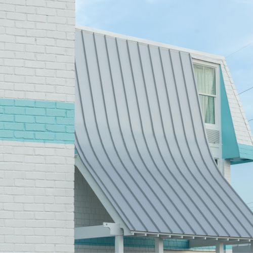 The image shows a modern building with a slanted metal roof, white brick walls, and a turquoise stripe. Sky in the background.