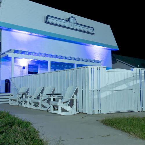 A building with blue lighting has a white fence and two chairs on the patio. It's nighttime and the exterior is well-lit.