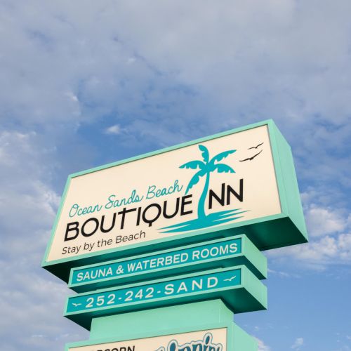 A sign for "Ocean Sands Beach Boutique Inn" offering a sauna, cookies, and sips by the beach, with a contact number.