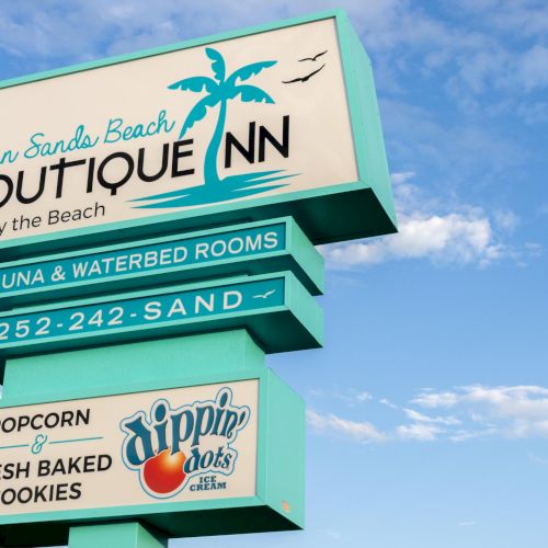 Sign for Ocean Sands Beach Boutique Inn with amenities listed like sauna and fresh baked cookies, set against a cloudy sky.