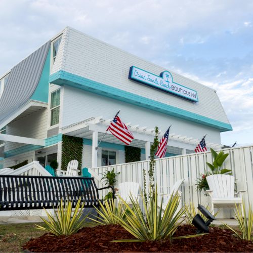 A white and turquoise building with flags, a bench, and plants in front, conveys a welcoming and stylish atmosphere.