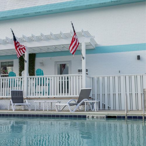 A pool in front of a boutique inn with American flags and lounge chairs, featuring a white and blue exterior.