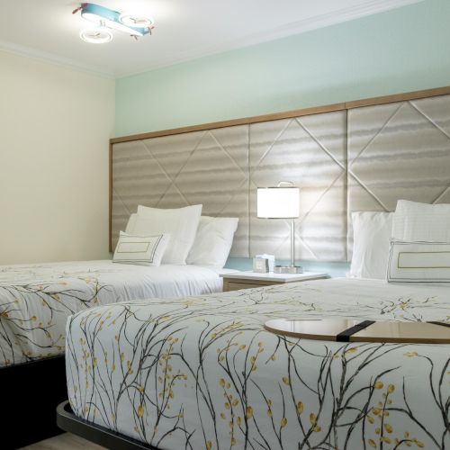 The image shows a hotel room with two neatly made beds, decorative headboards, elegant bedding, and a lamp on a nightstand between them.