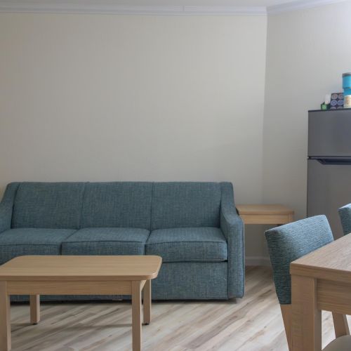 The image shows a living room with a blue sofa, wooden table, chairs, and a small fridge in the corner topped with items.