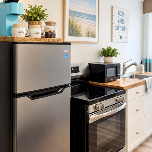 A bright kitchen corner with a stainless fridge, stove and microwave, plus mugs, plants, and wall art reading &ldquo;Good Vibes Stay Here.&rdquo;