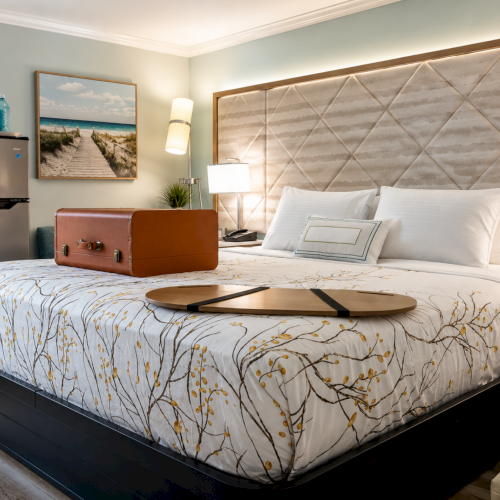A bright hotel bedroom with a patterned bedspread, tufted headboard, bedside lamps, and a mini fridge with bottled drinks.