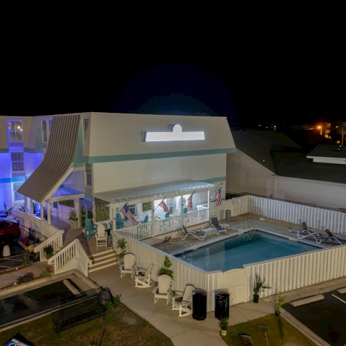 A nighttime view of a lit outdoor swimming pool area next to a building, with some seating and a grassy section in the foreground.