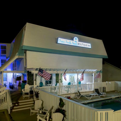 The image shows a building at night, adorned with a sign reading "Ocean Front Beach Boutique", surrounded by a pool and American flags.