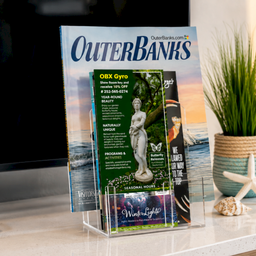 A stack of &ldquo;OuterBanks&rdquo; travel magazines on a desk near a lamp and potted grass, inside a magazine holder.