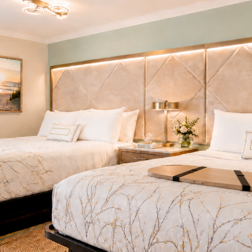 A bright, modern bedroom with two neatly made beds, decorative pillows, warm wall lighting, a framed beach photo, and a table vase of flowers.