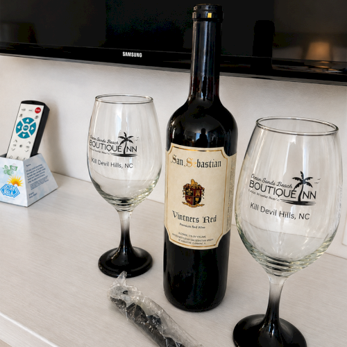 A dark wine bottle labeled &ldquo;San Sebastian&rdquo; sits between two branded wine glasses on a table, with a remote nearby.