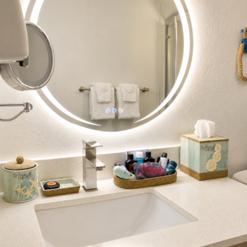 A modern bathroom vanity with a round illuminated mirror, two folded towels, and toiletries on the countertop.
