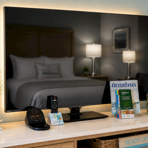 A flat-screen TV shows a bed scene, while a shelf beside it holds remotes, books, a plant, and seashell decor.