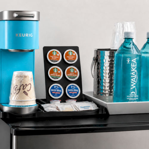 Blue Keurig with K-cup pods, Swiss Miss cocoa mix, and two Waiakea Hawaiian volcanic water bottles on the counter.