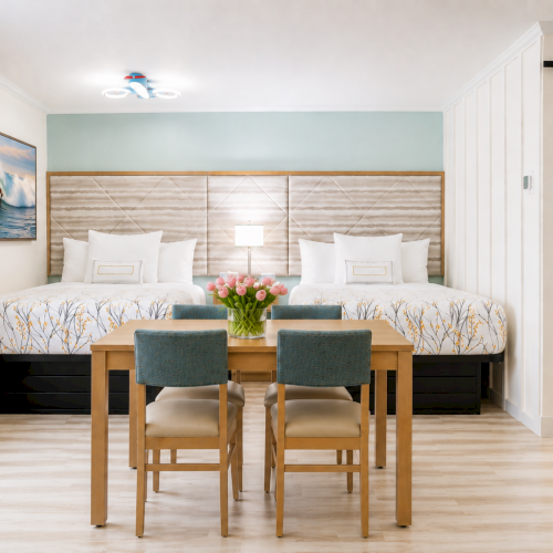 Bright hotel suite with two beds, ocean wall art, a wooden table, and a view into a modern bathroom.