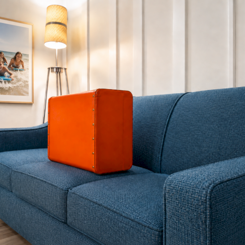 A cozy living room: blue sofa, orange suitcase-like trunk, floor lamp, and a framed beach photo on the wall.