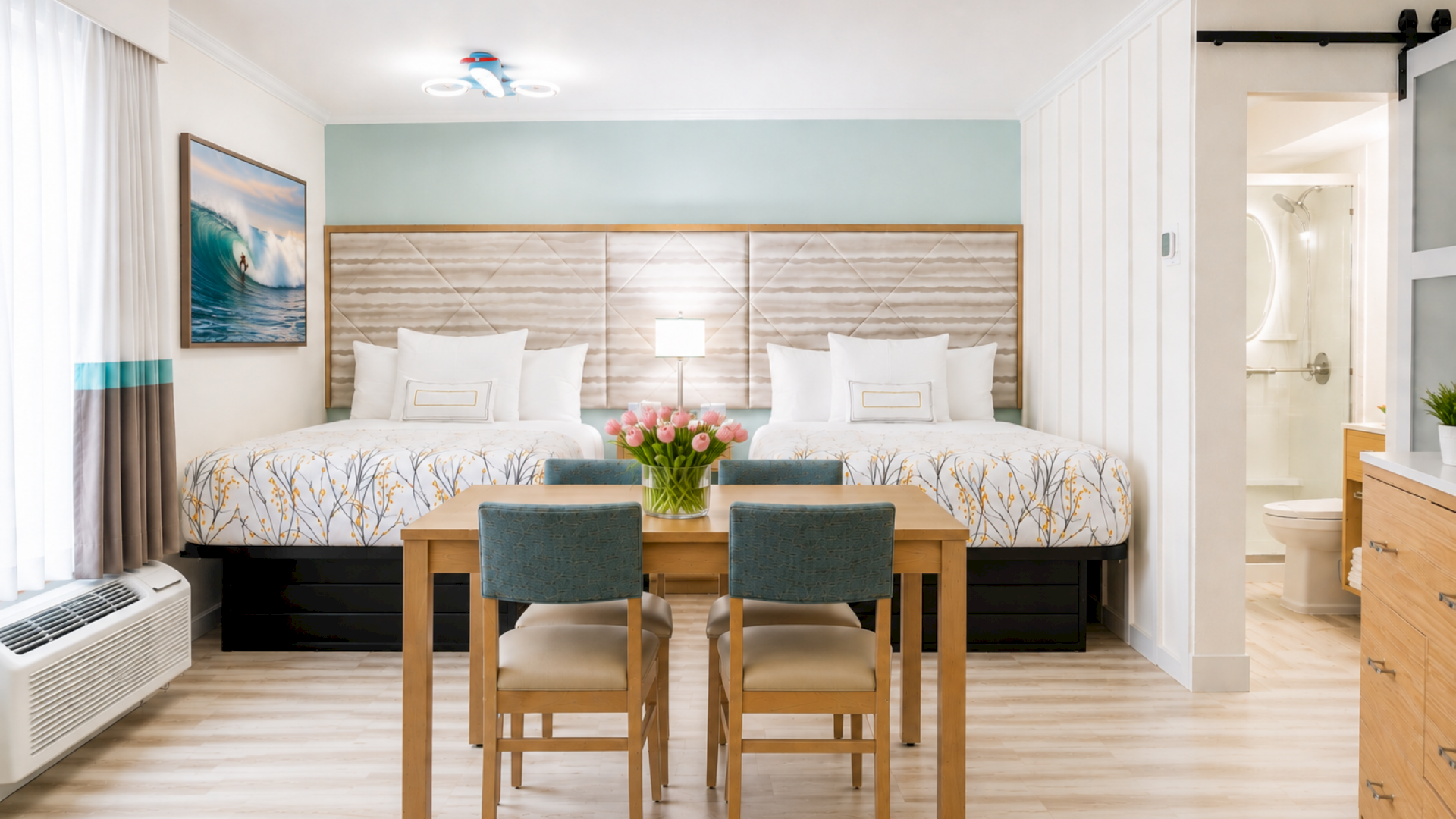 A bright hotel-style bedroom with two beds, a table with chairs, tulips in a vase, and an open ensuite bathroom doorway.