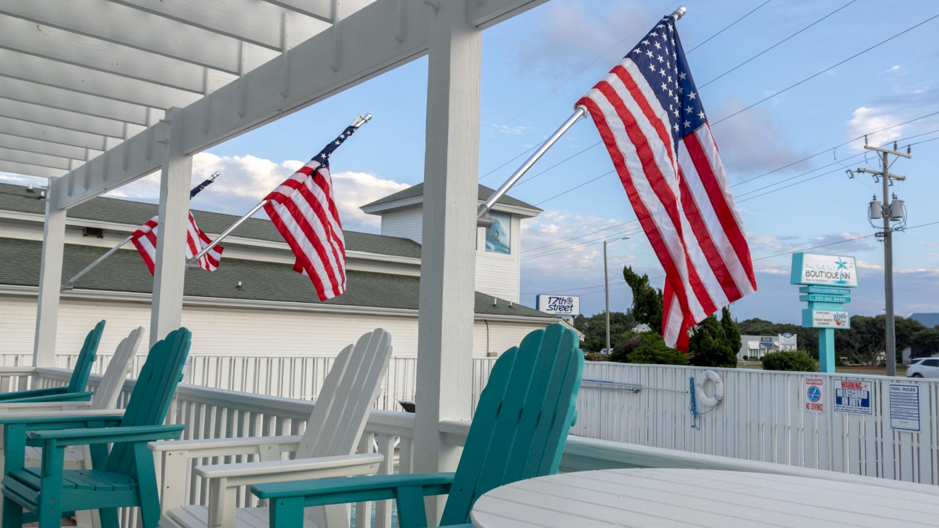 Outdoor seating area with teal chairs, American flags, and a white fence. A building and power lines are visible in the background.