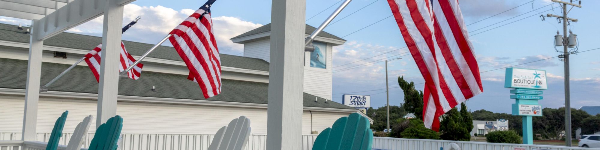Outdoor seating area with teal chairs, American flags, and a white fence. A building and power lines are visible in the background.