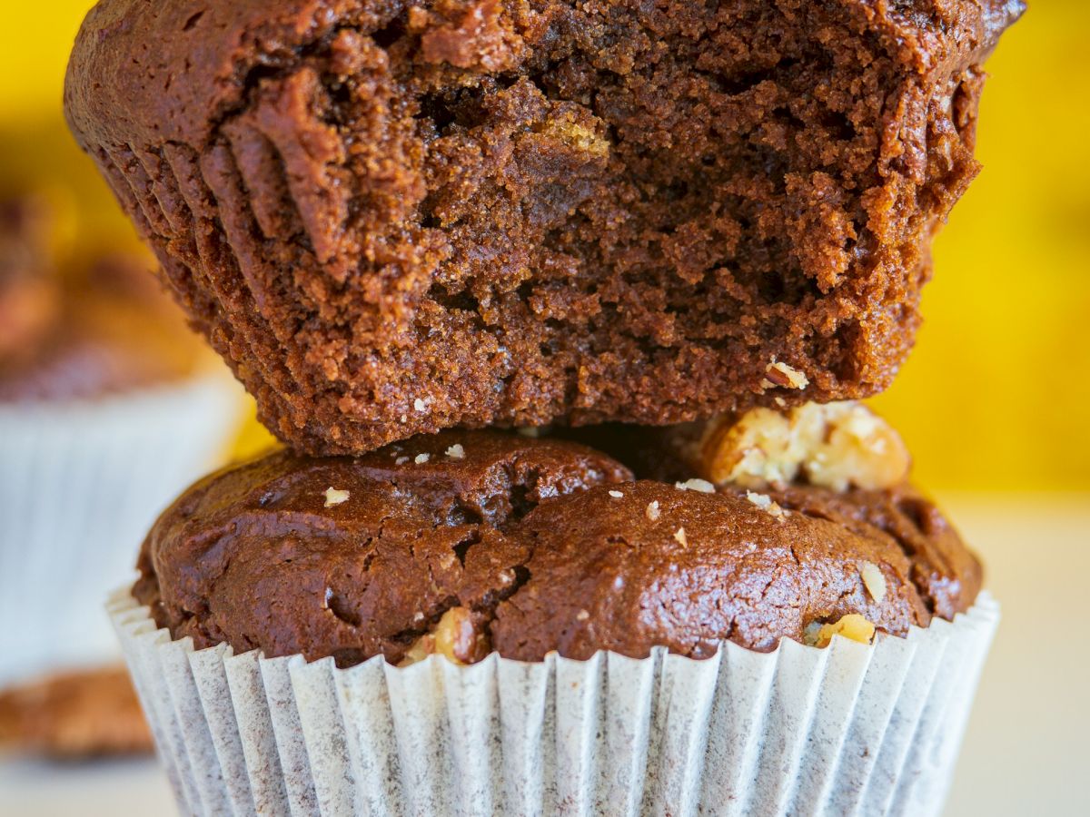 Close-up of a chocolate muffin split open, showing a fluffy dark crumb with walnut pieces against a bright yellow background.