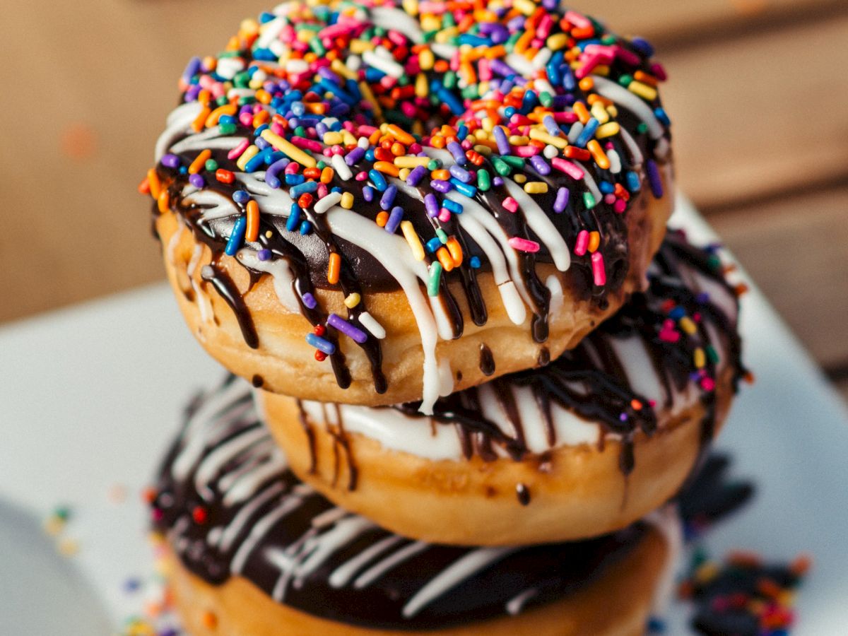 A colorful sprinkle-covered donut with chocolate and white icing drizzles, stacked on a tray.