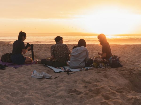 Friends sit on a sandy beach on blankets at sunset as waves roll in and birds fly overhead.