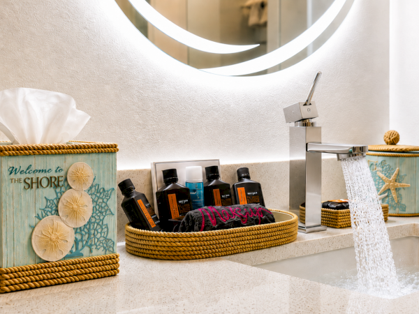 A bright bathroom sink with a waterfall faucet, soap and lotion bottles, a loofah/towel, and beach-themed d&eacute;cor on the counter.