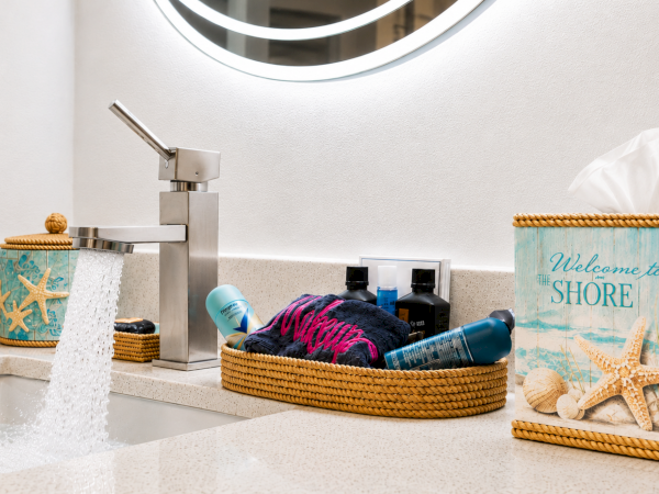 Bathroom sink with a running faucet, beach-themed toiletries, woven baskets, and a &ldquo;Welcome to the Shore&rdquo; tissue box.