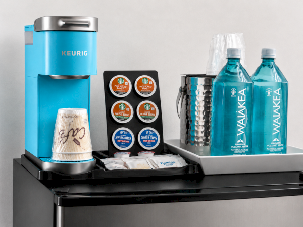 A blue Keurig coffee maker with assorted pods, Swiss Miss, and several blue Hawaiian volcanic water bottles.