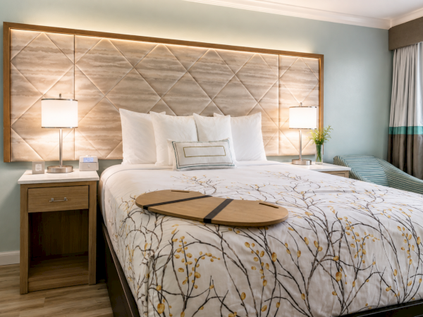 A modern hotel-style bedroom with a neatly made bed, bedside lamps, and geometric wall art mirrored behind it.