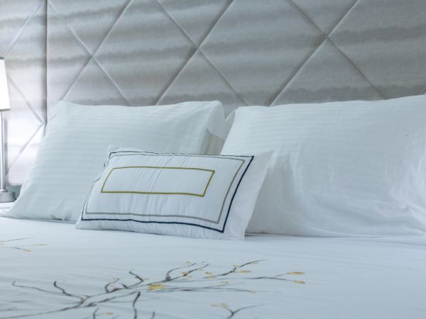 A neatly made bedroom bed with white sheets and pillows, a decorative cushion, and a bedside lamp against a padded headboard.