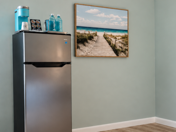 A stainless fridge in a clean room, with bottled water and a coffee/tea dispenser on top, plus a framed beach walkway photo.
