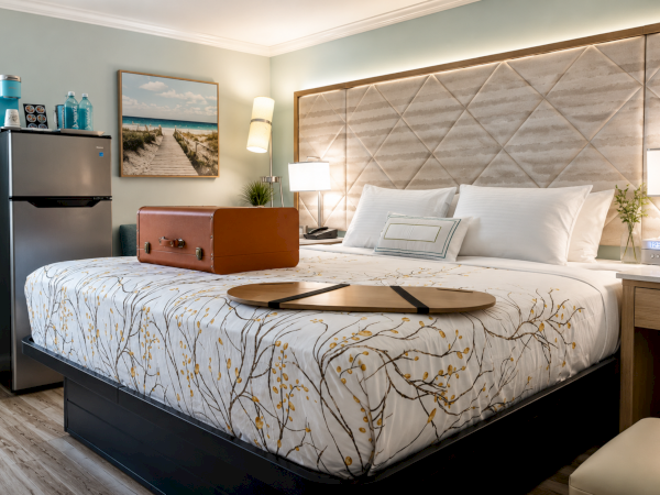 A modern hotel room with a neatly made bed, pillows, a suitcase, fridge, and bedside lamps, set beside a patterned headboard.