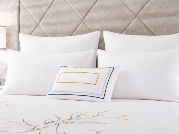 A hotel bed with white pillows, a decorative cushion, and a bedside lamp beside a tufted upholstered headboard.