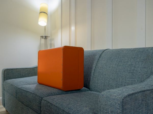 An orange suitcase sits on a blue upholstered couch under a wall lamp in a bright modern room.