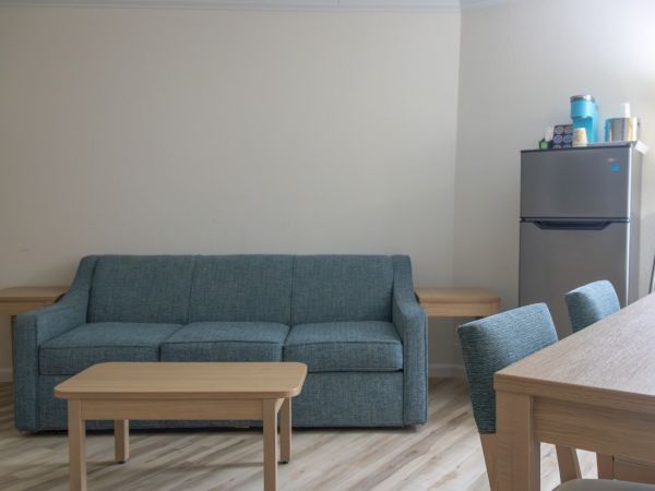 A bright living room with a blue sofa, wooden tables and chairs, and a refrigerator with pantry items in the corner.