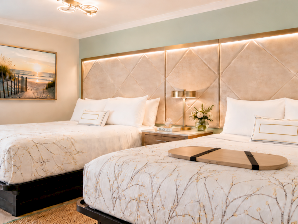 A bright modern bedroom with two twin beds, gold tufted headboard lighting, and beach-themed decor on the walls.