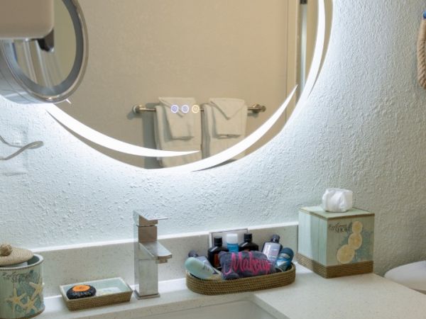 A bright bathroom vanity with a white sink, toiletries in baskets, and a round illuminated mirror reflecting the room.