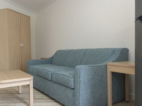 A small living room with a blue fabric sofa, a light wooden side table, a closet, and neutral walls under ceiling lights.