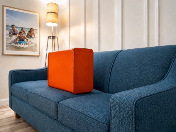 A cozy living room: blue sofa, orange trunk-like suitcase, standing lamp, and a framed beach photo on the wall.