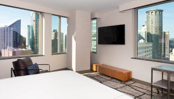 A modern hotel room with a bed, chair, TV, and views of a cityscape through large windows, featuring tall buildings outside.