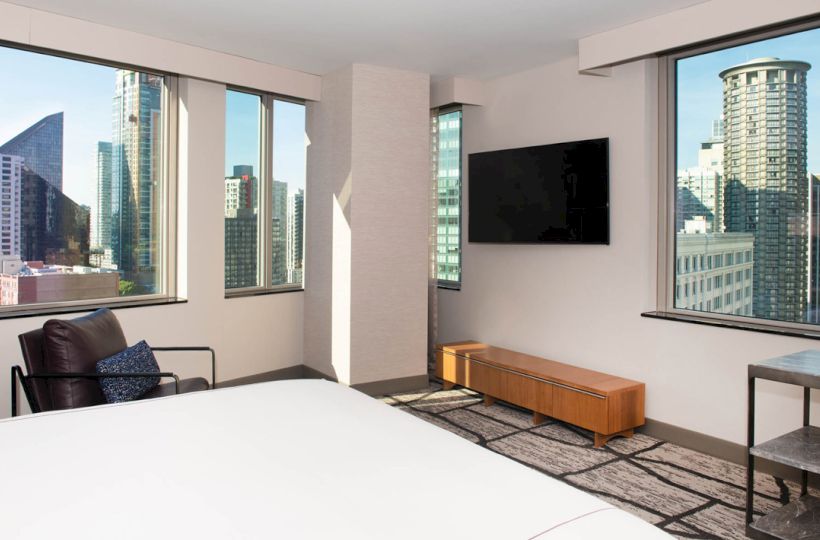 A modern hotel room with a bed, chair, TV, and views of a cityscape through large windows, featuring tall buildings outside.