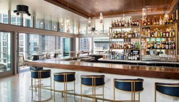 A modern bar with a wooden counter, stylish stools, and a fully stocked liquor shelf, surrounded by large windows.