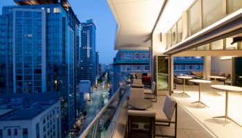 A modern balcony with tables and chairs overlooks a cityscape with high-rise buildings at dusk.