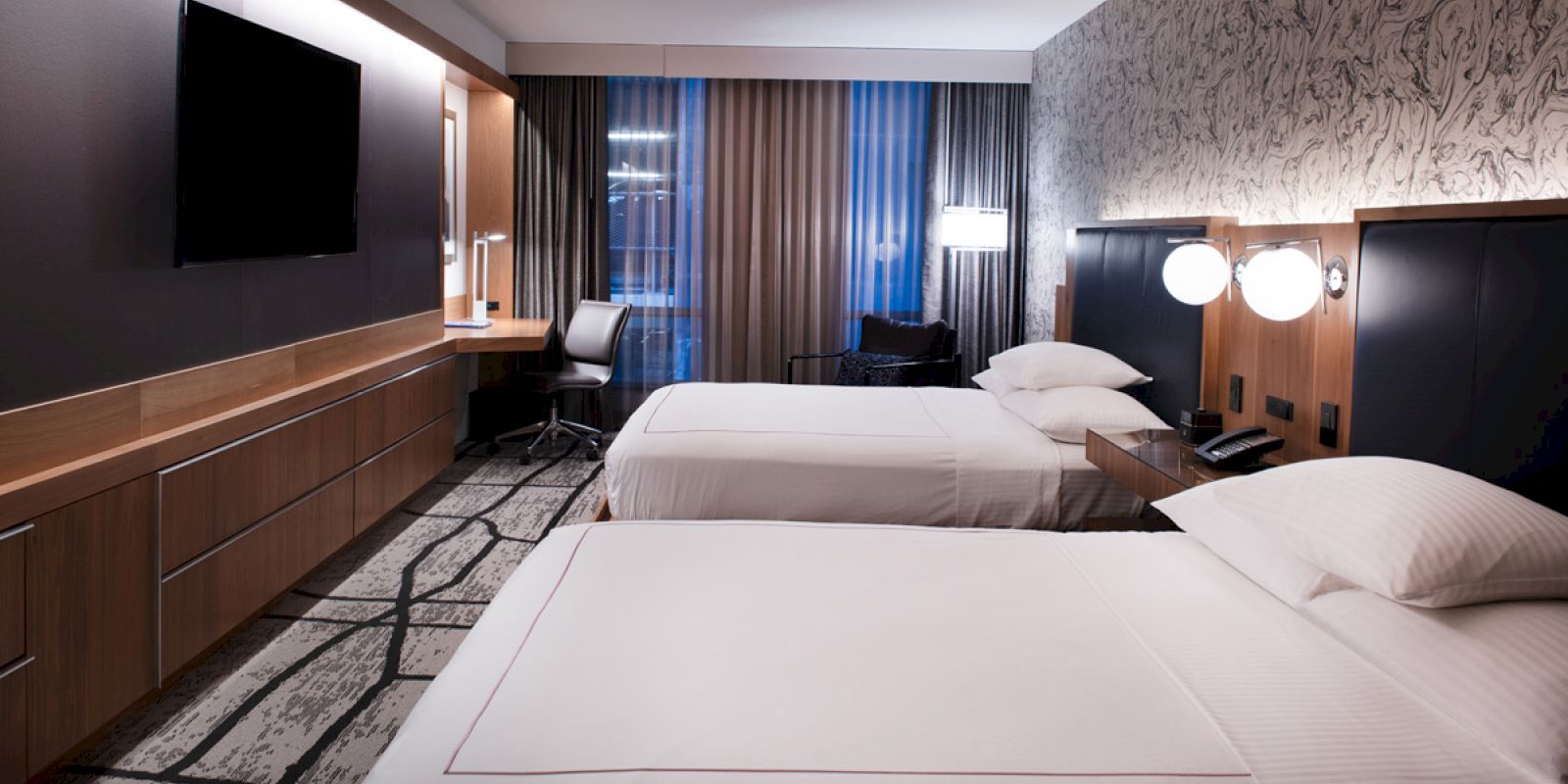 A modern hotel room with two beds, a large TV, a desk, and contemporary decor.