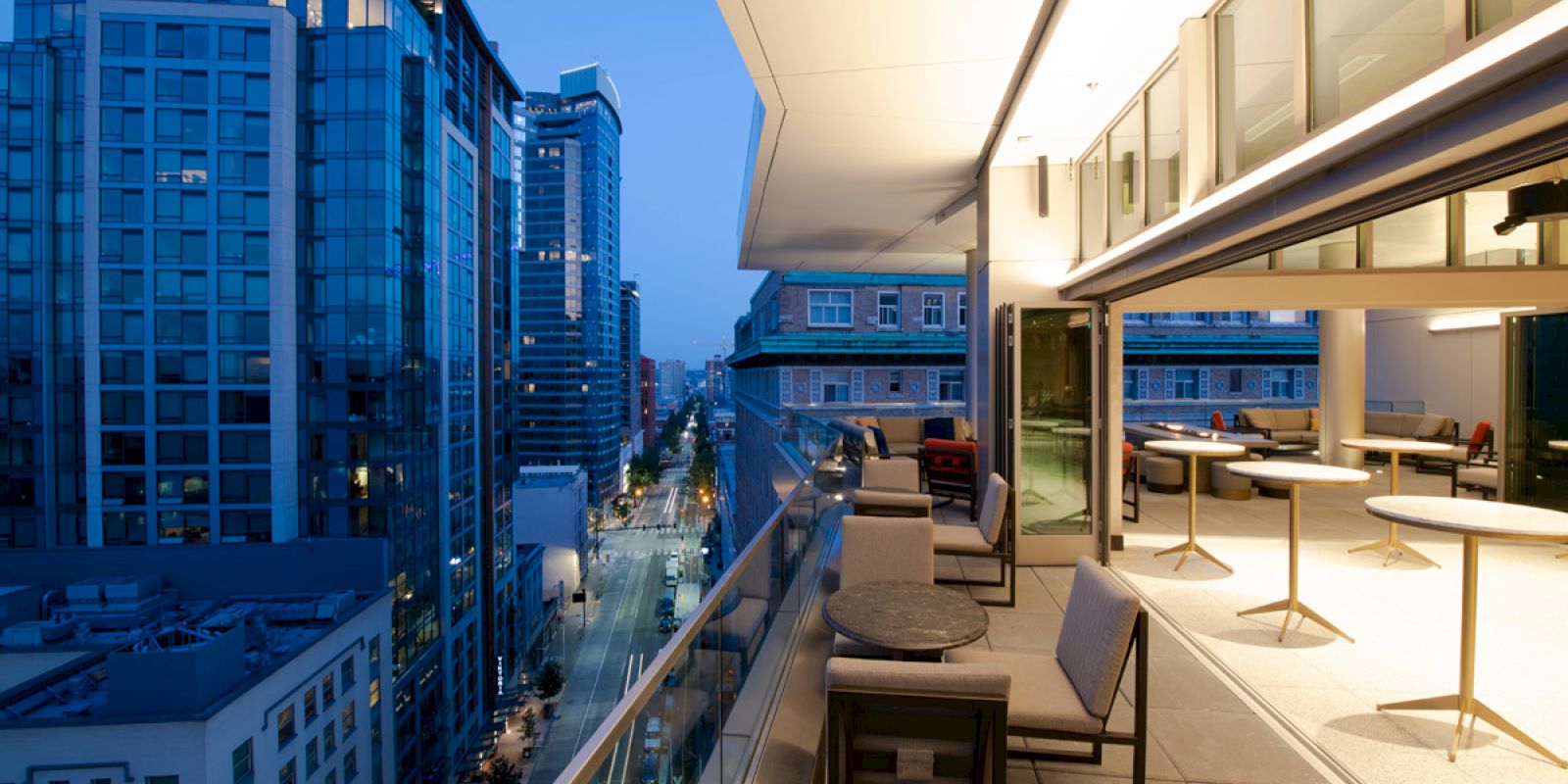A modern rooftop terrace with outdoor seating overlooks a cityscape of high-rise buildings at dusk, with a view of a street below.