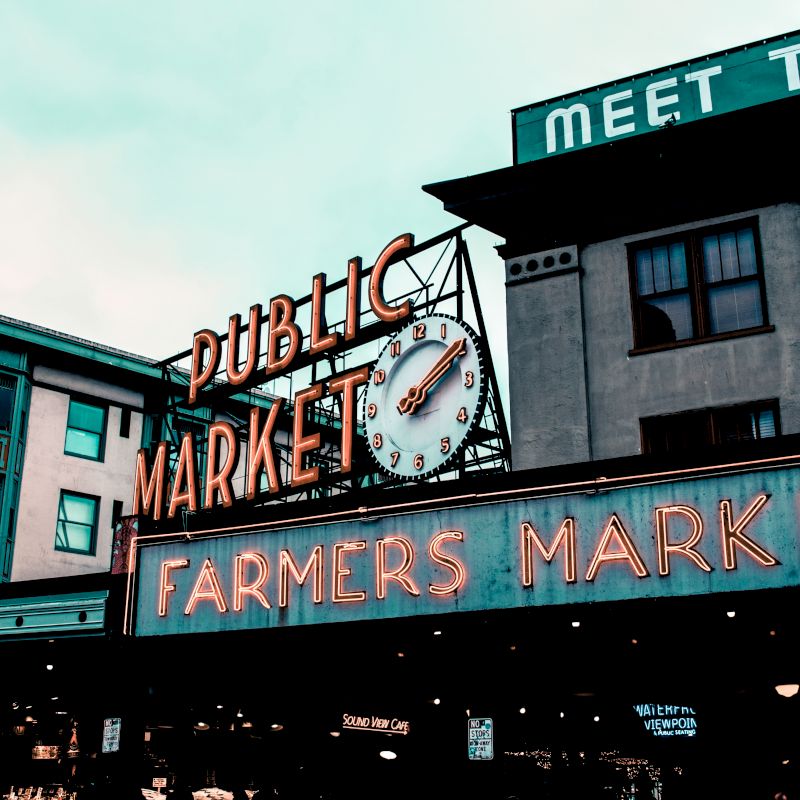 The image shows a public market with a neon sign that reads "Farmers Market." There's a large clock above the entrance.